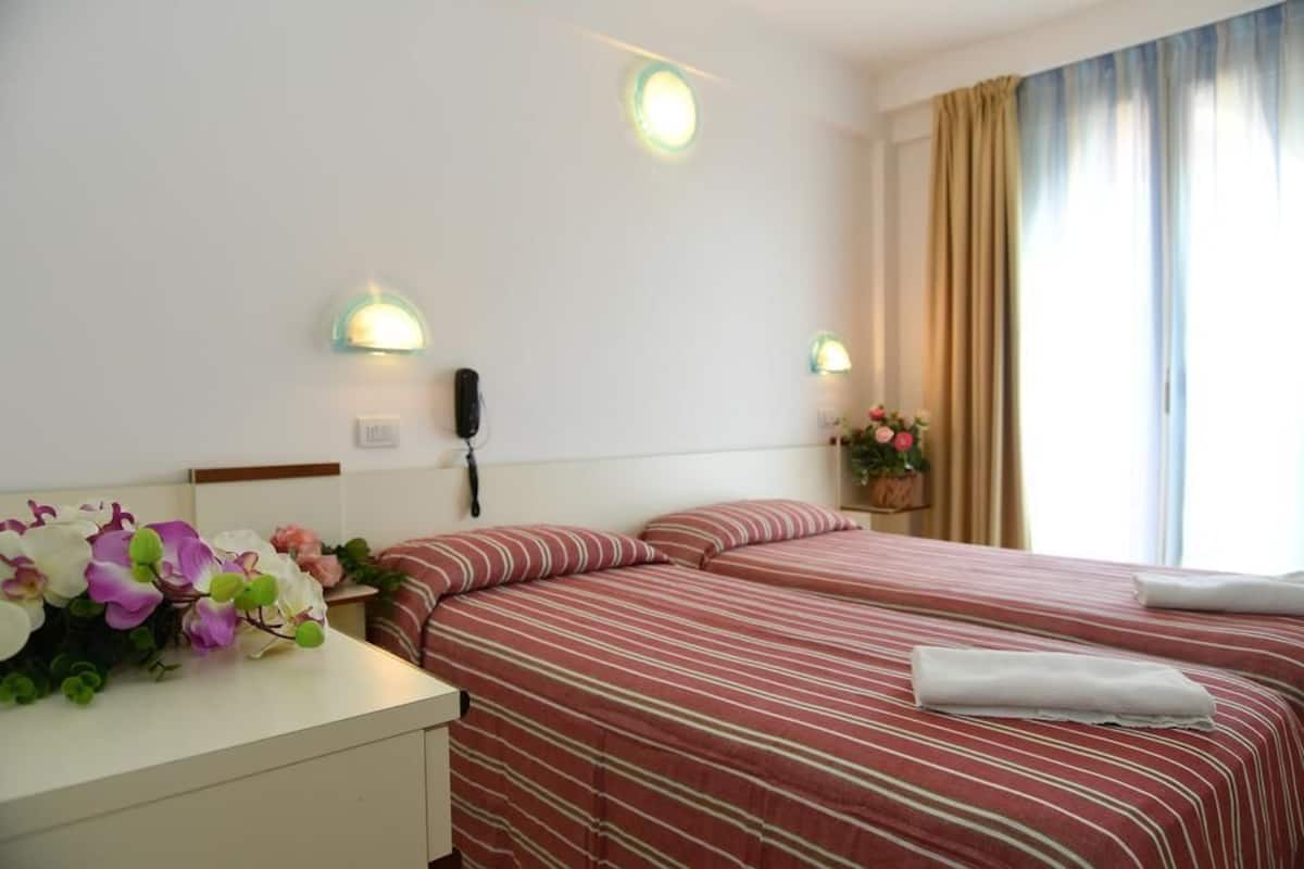 A hotel room with two beds, a bedside table with a lamp, and a window with curtains.