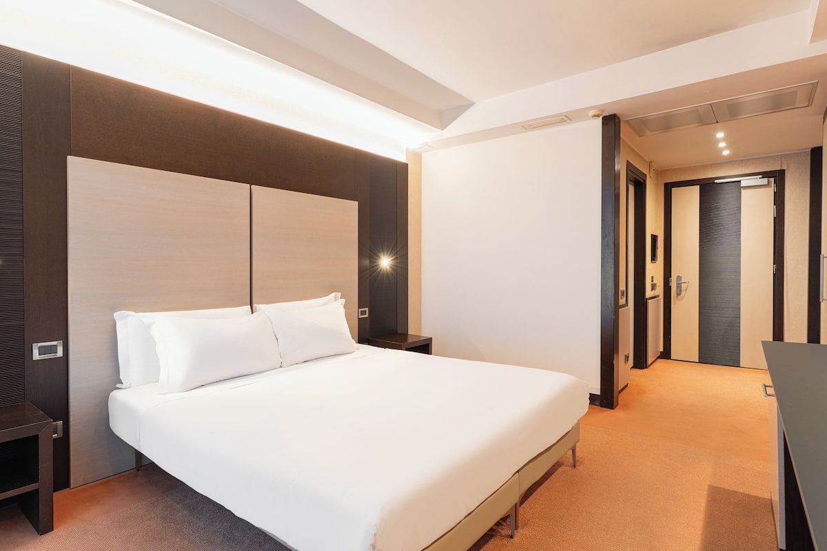 A modern hotel room with a large bed, a bedside table, a wardrobe, and a door leading to another room.