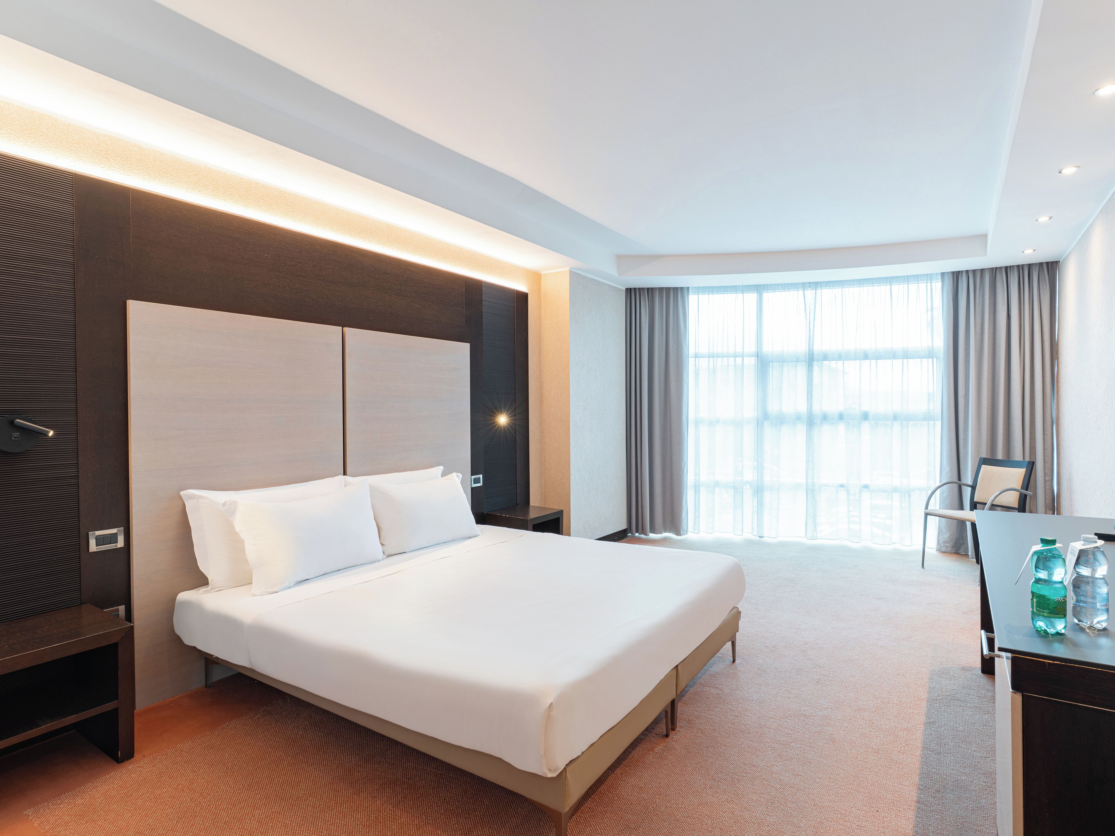 Ein modernes Hotelzimmer mit einem großen Bett, einem Schreibtisch mit Stuhl und einem großen Fenster mit Vorhängen.