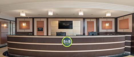 Una recepción con el logotipo de 'B&B Hotels', un televisor de pantalla plana y un área de recepción de diseño moderno.