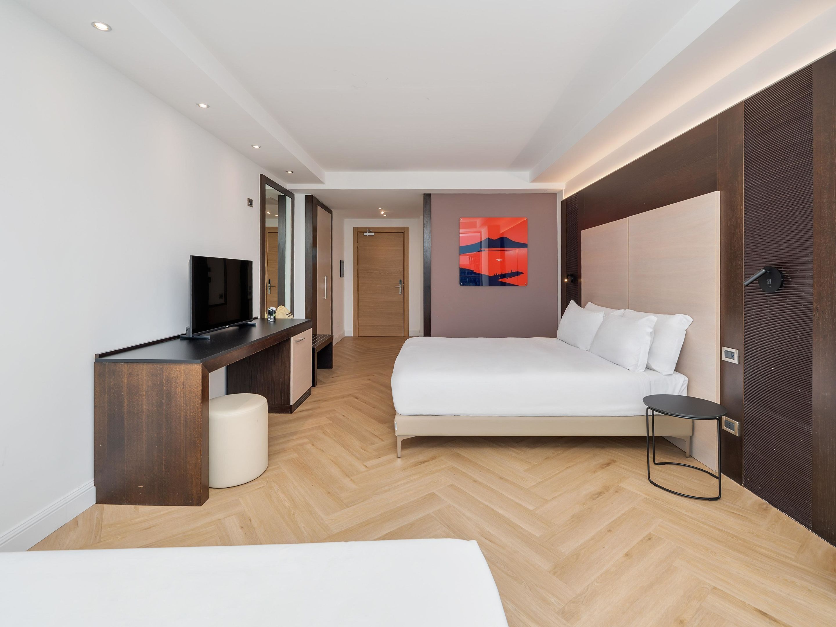 Una habitación de hotel moderna con cama, escritorio con televisor, mesita de noche y un cuadro abstracto grande en la pared.