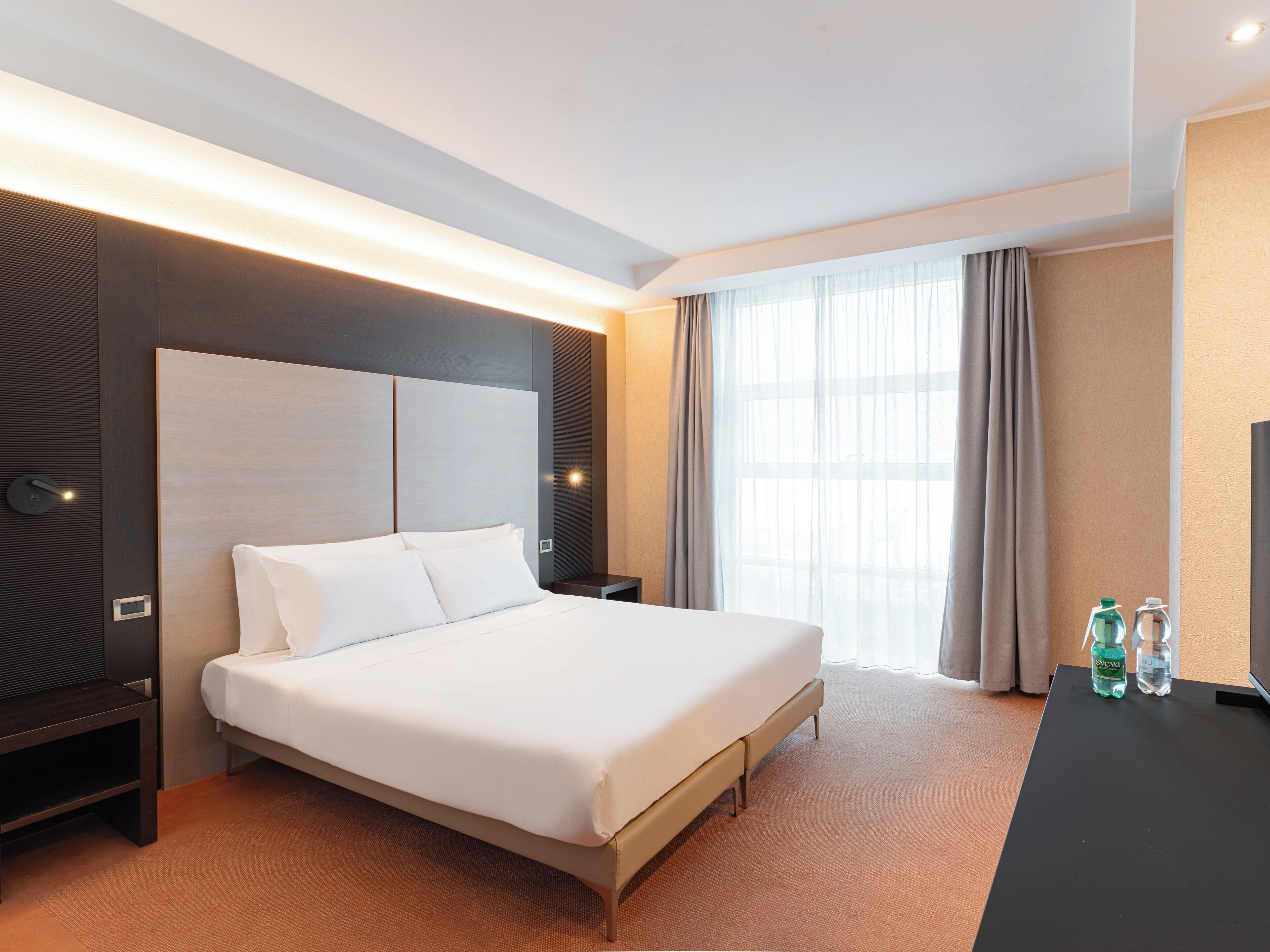 Habitación de hotel moderna con una cama grande, una mesita de noche, un televisor de pantalla plana y un ventanal con cortinas.
