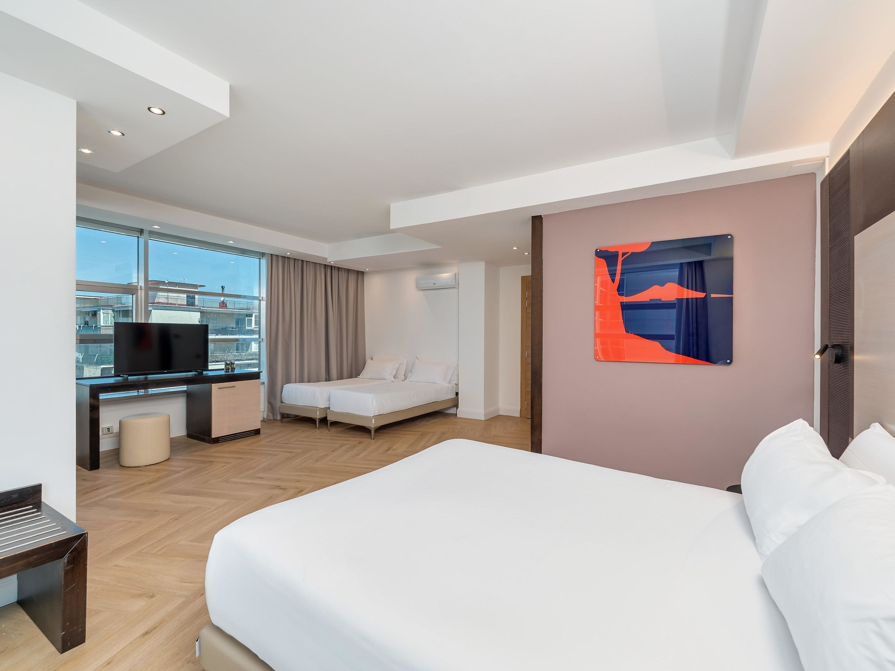 Une chambre d’hôtel moderne dotée d’un grand lit, d’un bureau avec une télévision, d’un petit coin salon et d’une grande fenêtre donnant sur la ville.