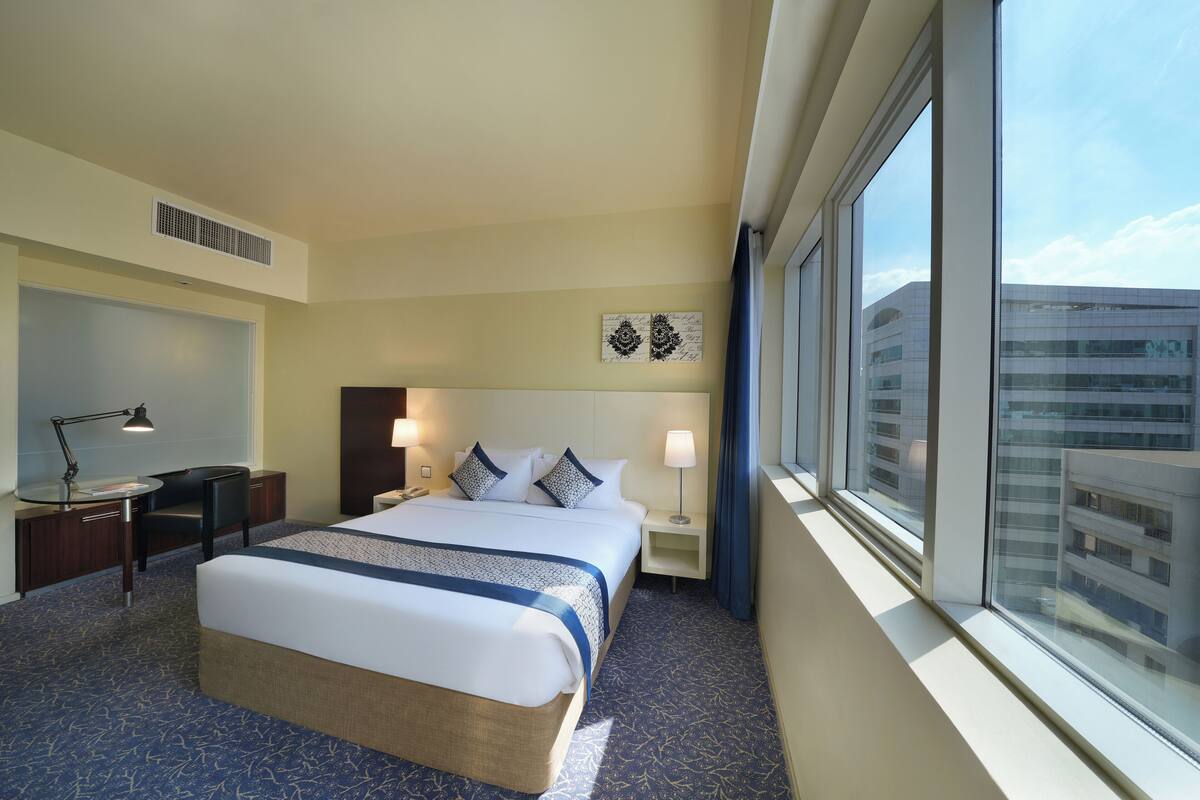 Une chambre d’hôtel avec un grand lit, un bureau, une chaise, une table de chevet, une lampe, une fenêtre donnant sur la ville et une bouche d’aération au plafond.