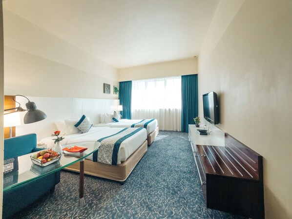 A hotel room with two beds, a desk, a TV, and a glass coffee table with fruit.