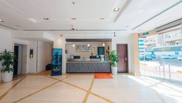 A hotel reception area with a modern design, featuring a reception desk, potted plants, and a view of the street outside.
