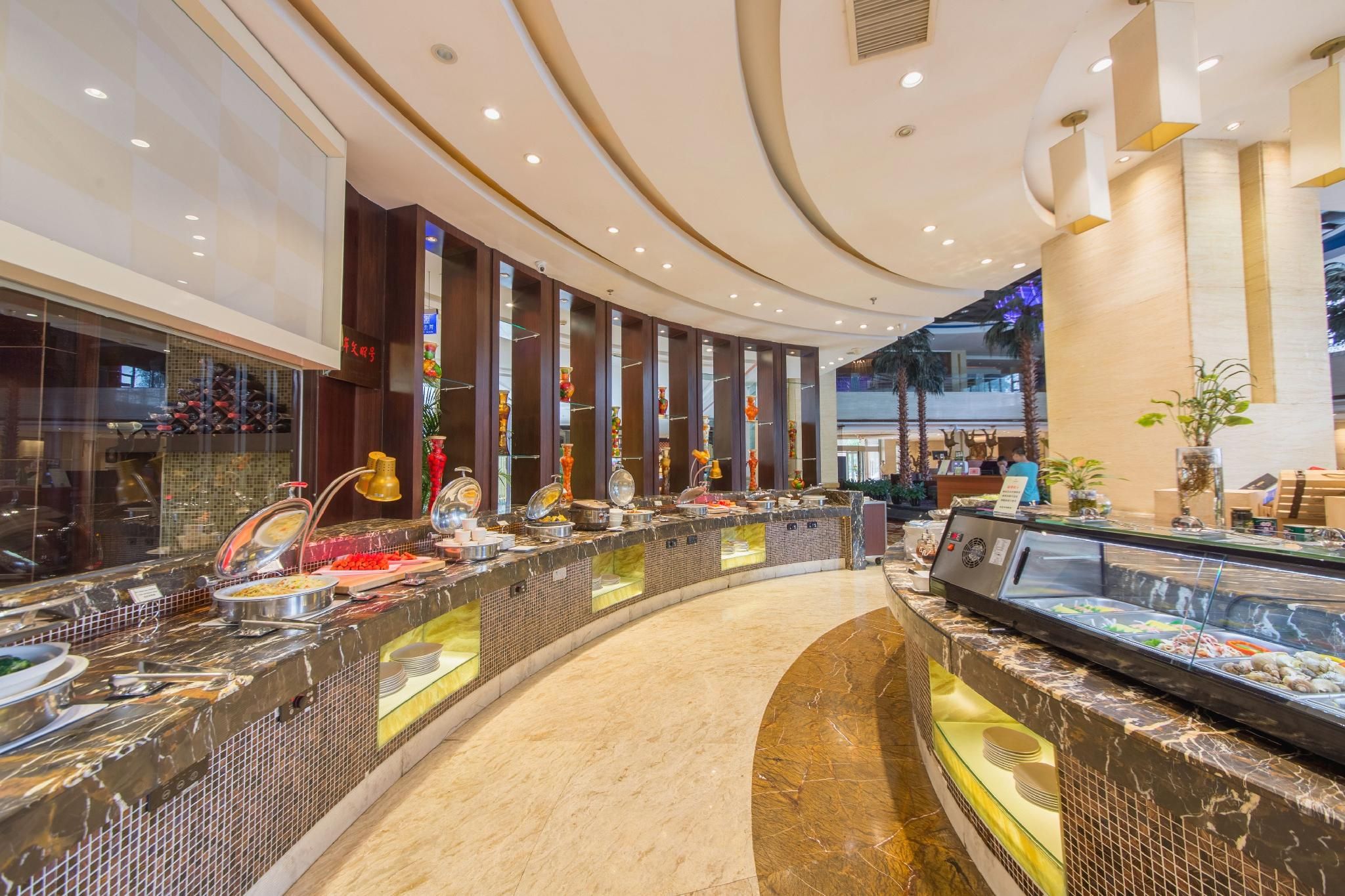 Un hall de hotel con un mostrador de recepción curvo, una vitrina con productos alimenticios y una zona de estar con vista al hall.