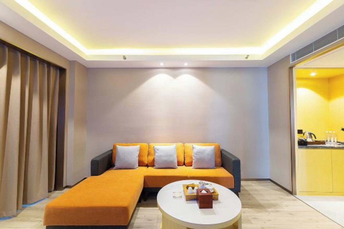 Una habitación de hotel moderna con un sofá seccional, una mesa de centro redonda y una cocina visible a través de un umbral abierto.