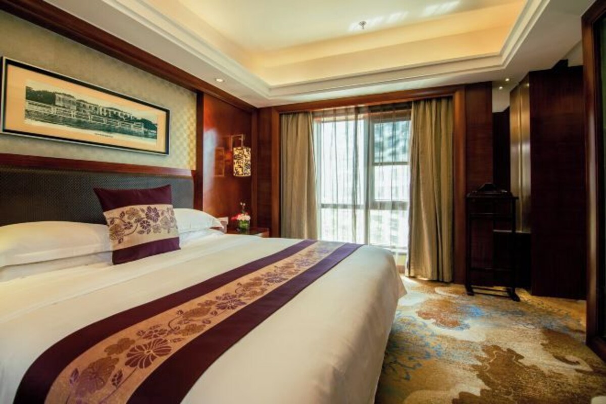 Una habitación de hotel con una cama grande, una mesita de noche con lámpara, un cuadro enmarcado en la pared y una ventana con cortinas.