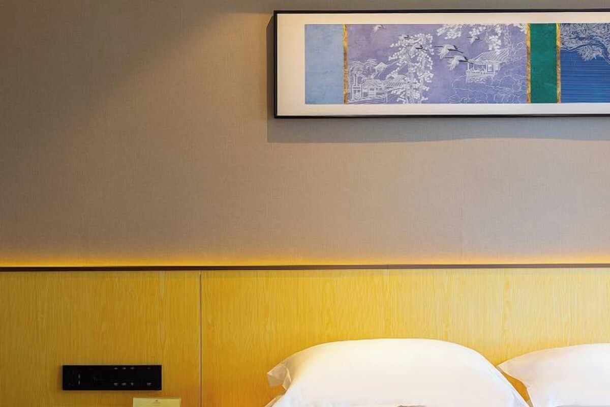 A hotel room with a wooden headboard, a framed artwork above the bed, and a control panel on the wall.