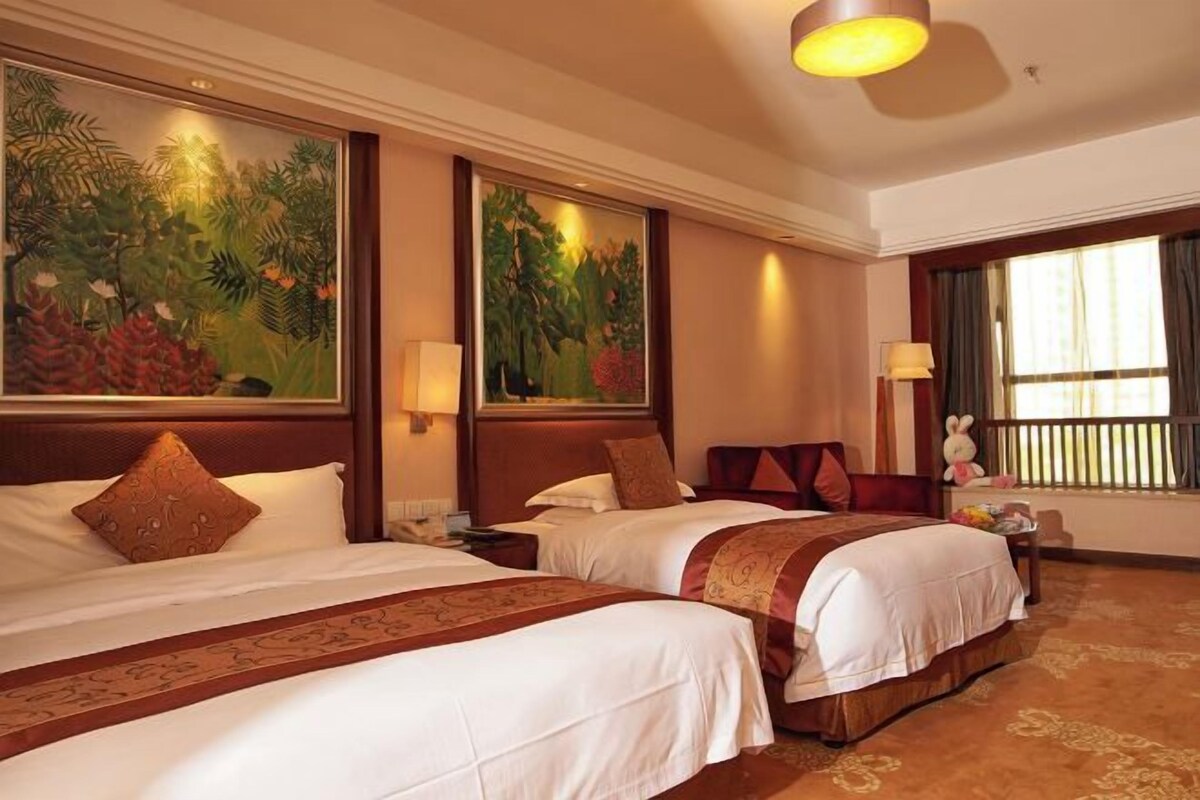 A hotel room with two beds, a large painting on the wall, a window with curtains, and a nightstand with a lamp.