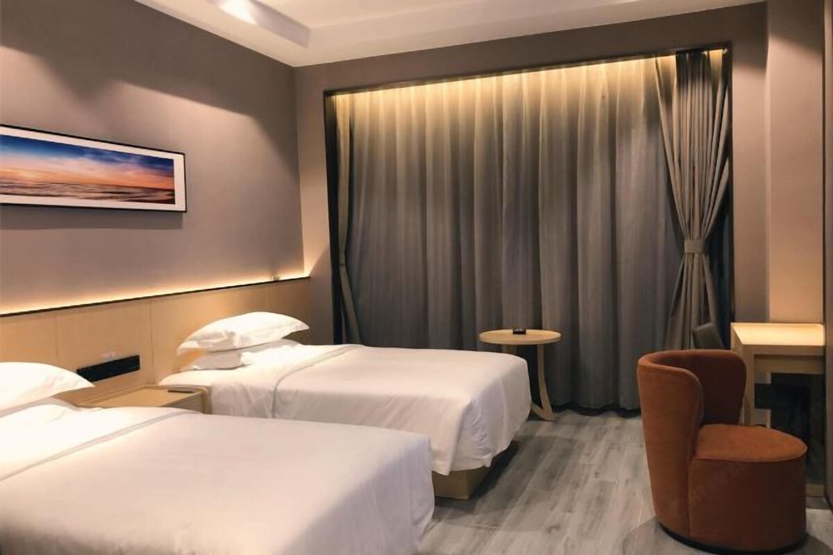 Habitación de hotel con dos camas, una mesita redonda pequeña, una silla, un cuadro en la pared y un ventanal grande con cortinas.
