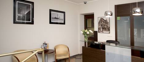 A reception area with a glass desk, two wicker chairs, a small table with a bottle, and framed pictures on the wall.