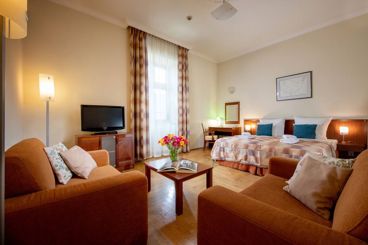 A hotel room with two sofas, a TV, a desk, a bed, and a painting on the wall.