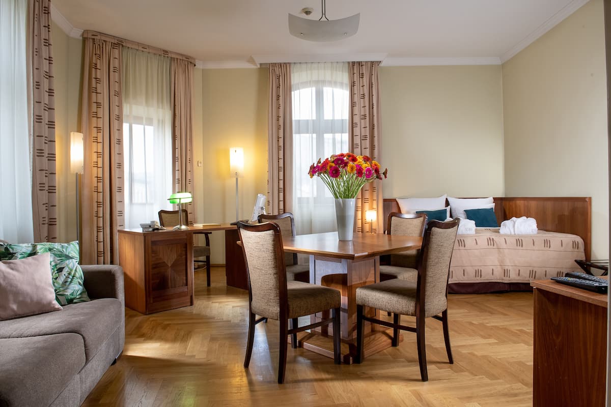 A hotel room with a dining table, chairs, a sofa, a bed, and a vase of flowers.