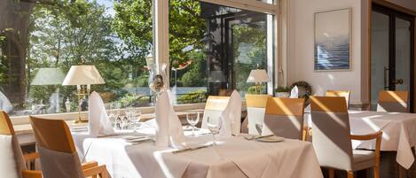 A dining area with tables set for service, large windows, and a view of greenery outside.