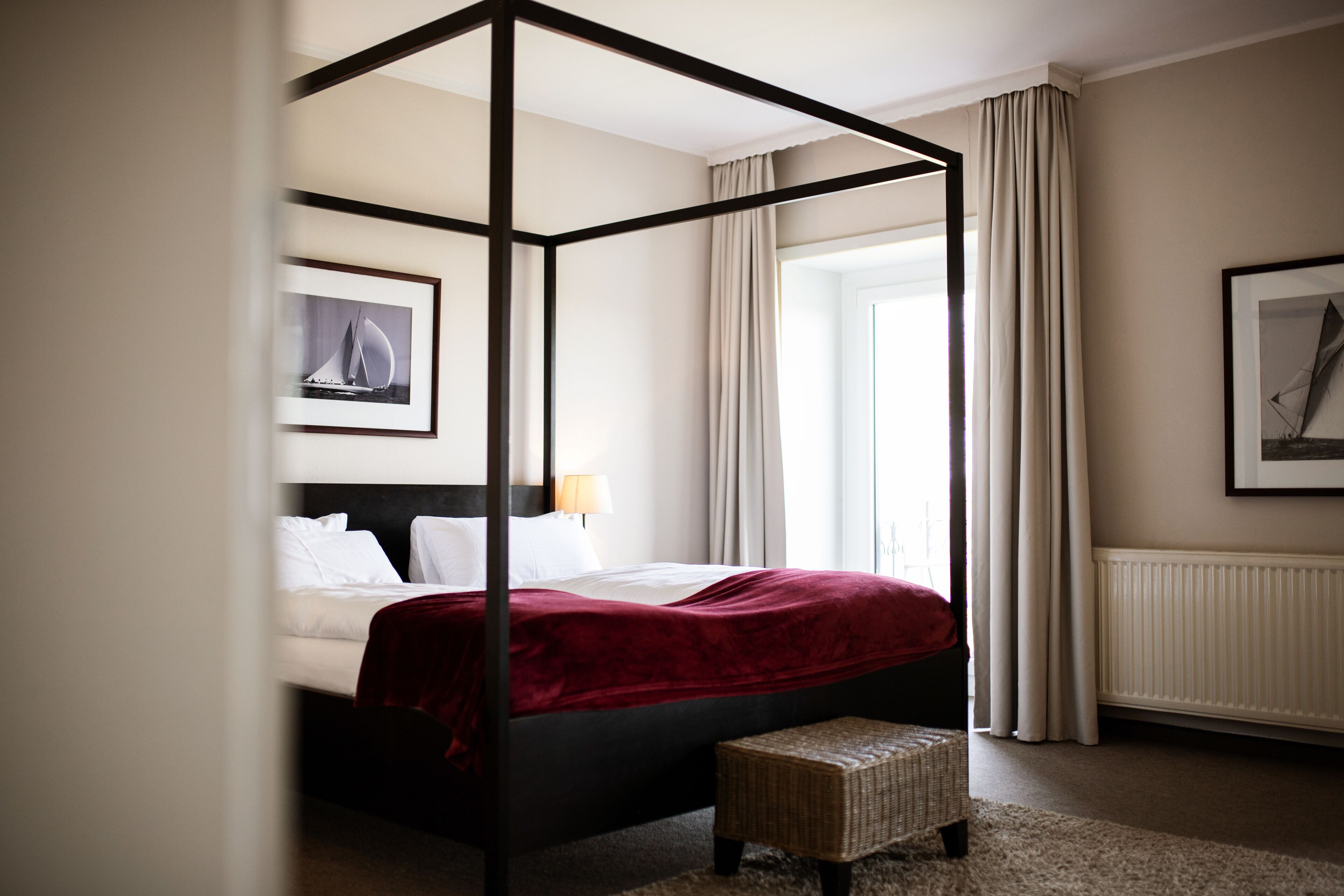A four-poster bed with a red bedspread, a wicker bench, and two framed pictures on the walls.