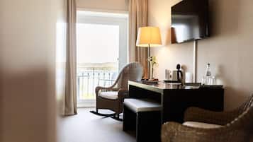 Une chambre d’hôtel équipée d’un bureau, de deux fauteuils en osier, d’un téléviseur fixé au mur, d’une lampe et offrant une vue sur l’extérieur grâce à une grande fenêtre.