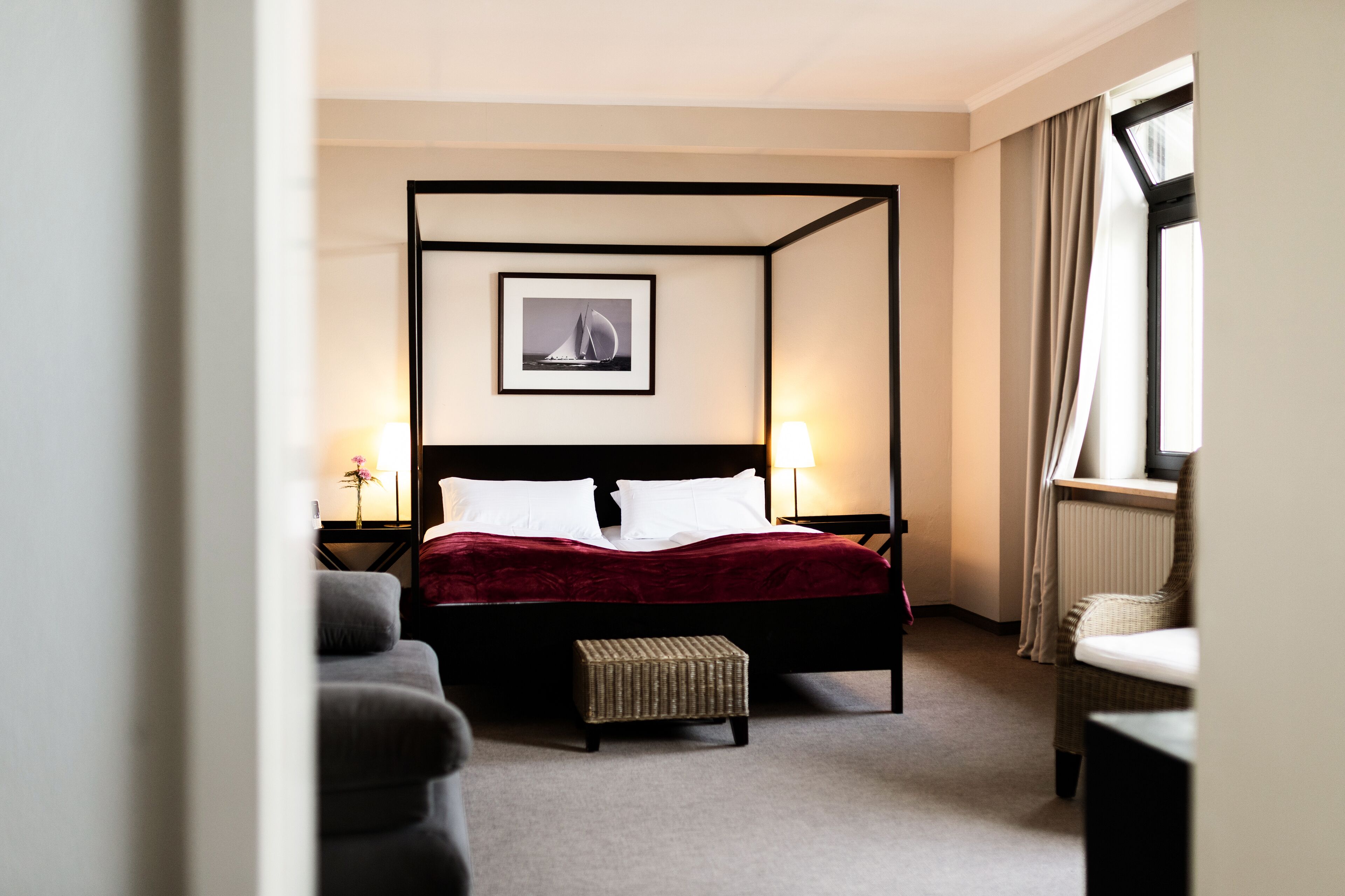 A four-poster bed with a red bedspread, a framed picture above it, and a window with curtains to the right.
