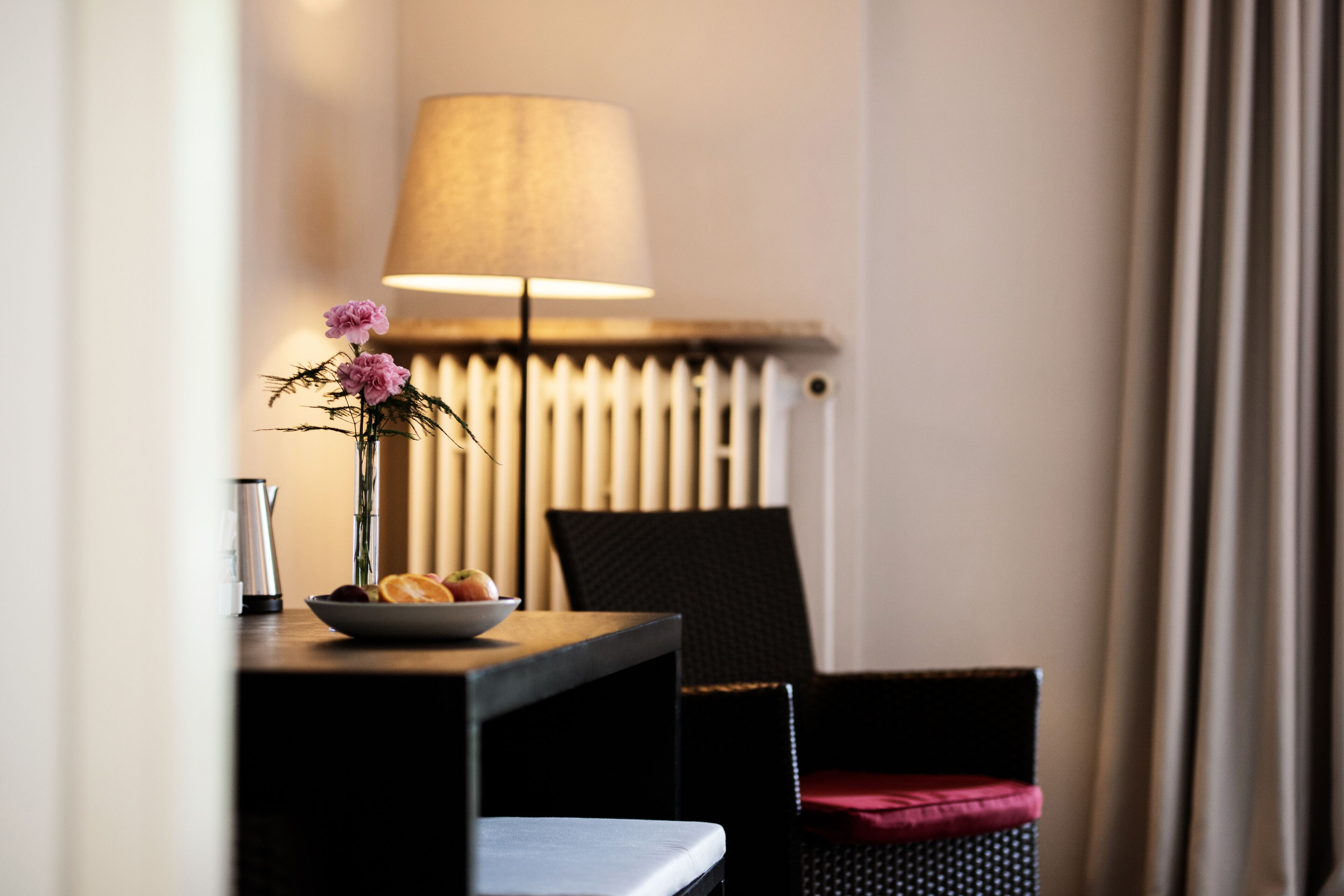 A hotel room with a table, a lamp, a chair, and a radiator.