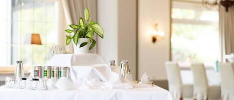 A dining table set for a meal with a white tablecloth, a potted plant, and various dining items.