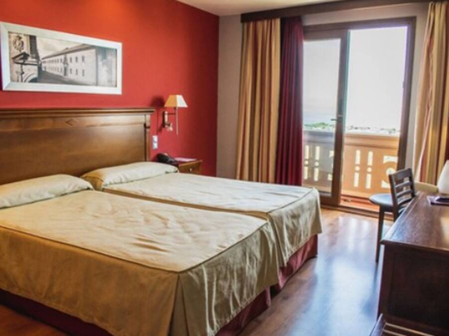 A hotel room with two beds, a desk, a chair, a window with curtains, and a painting on the wall.