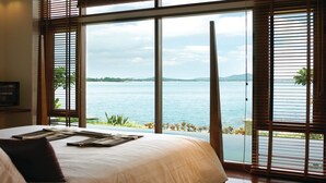A bedroom with a large window overlooking a body of water, a bed with a white comforter, and wooden blinds. - The Sarann Koh Samui, Suites & Villas (Koh Samui)