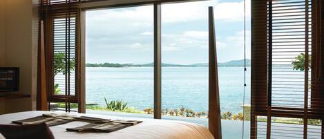 A bedroom with a large window overlooking a body of water, a bed with a white comforter, and wooden blinds.