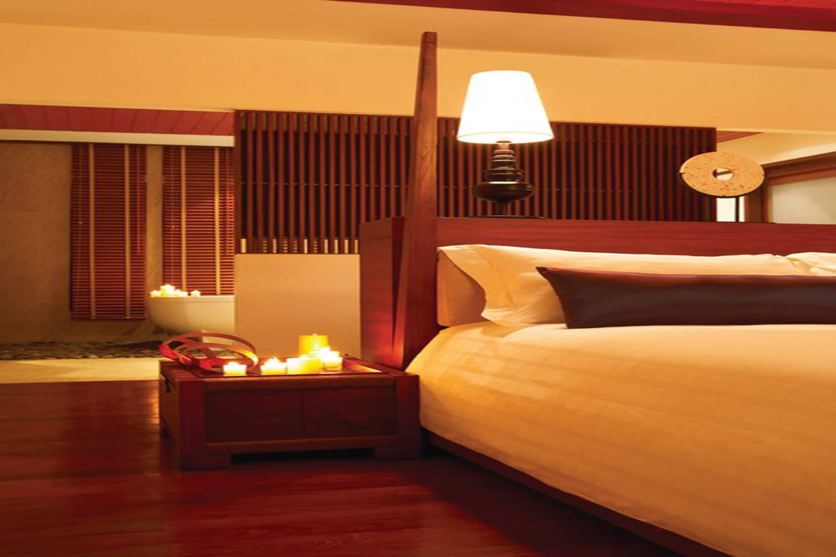 A hotel room with a wooden bed, a nightstand with candles, a wall clock, and a window with blinds.