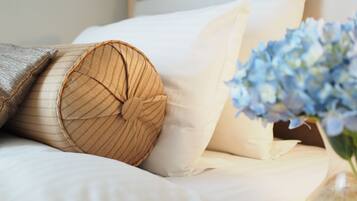 A neatly made bed with white linens, a rolled-up beige pillow, and a vase with blue flowers on a bedside table.