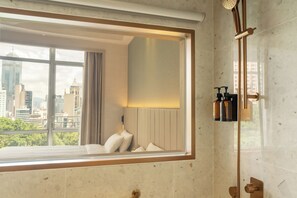 Casa de banho moderna com uma janela grande, chuveiro e uma cama vista para a cidade.