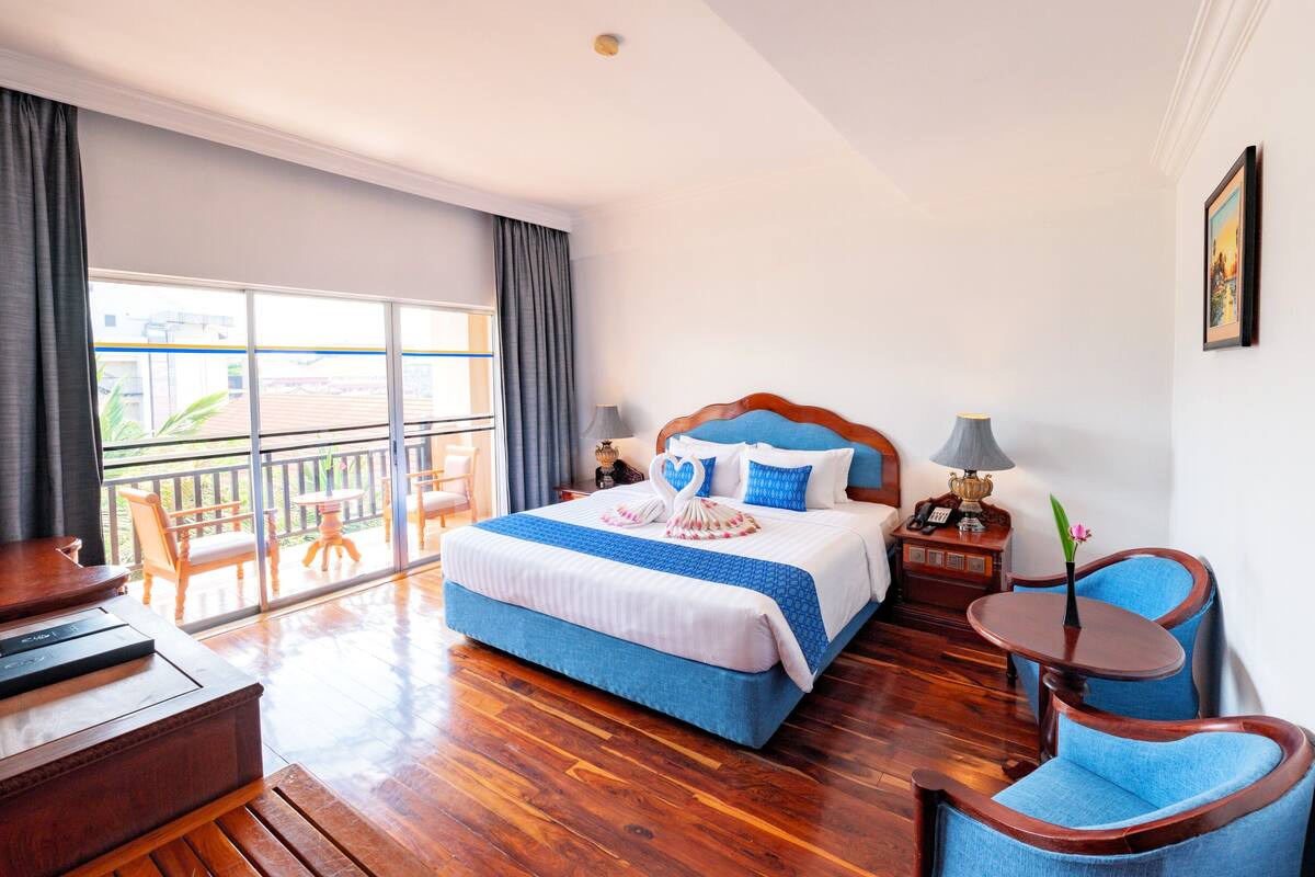 A hotel room with a large bed, a wooden floor, a balcony with a view, a desk, and a chair.