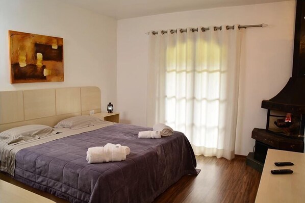 Una cama bien tendida con una colcha oscura y dos toallas enrolladas, un cuadro en la pared, una ventana con cortinas translúcidas y una chimenea.