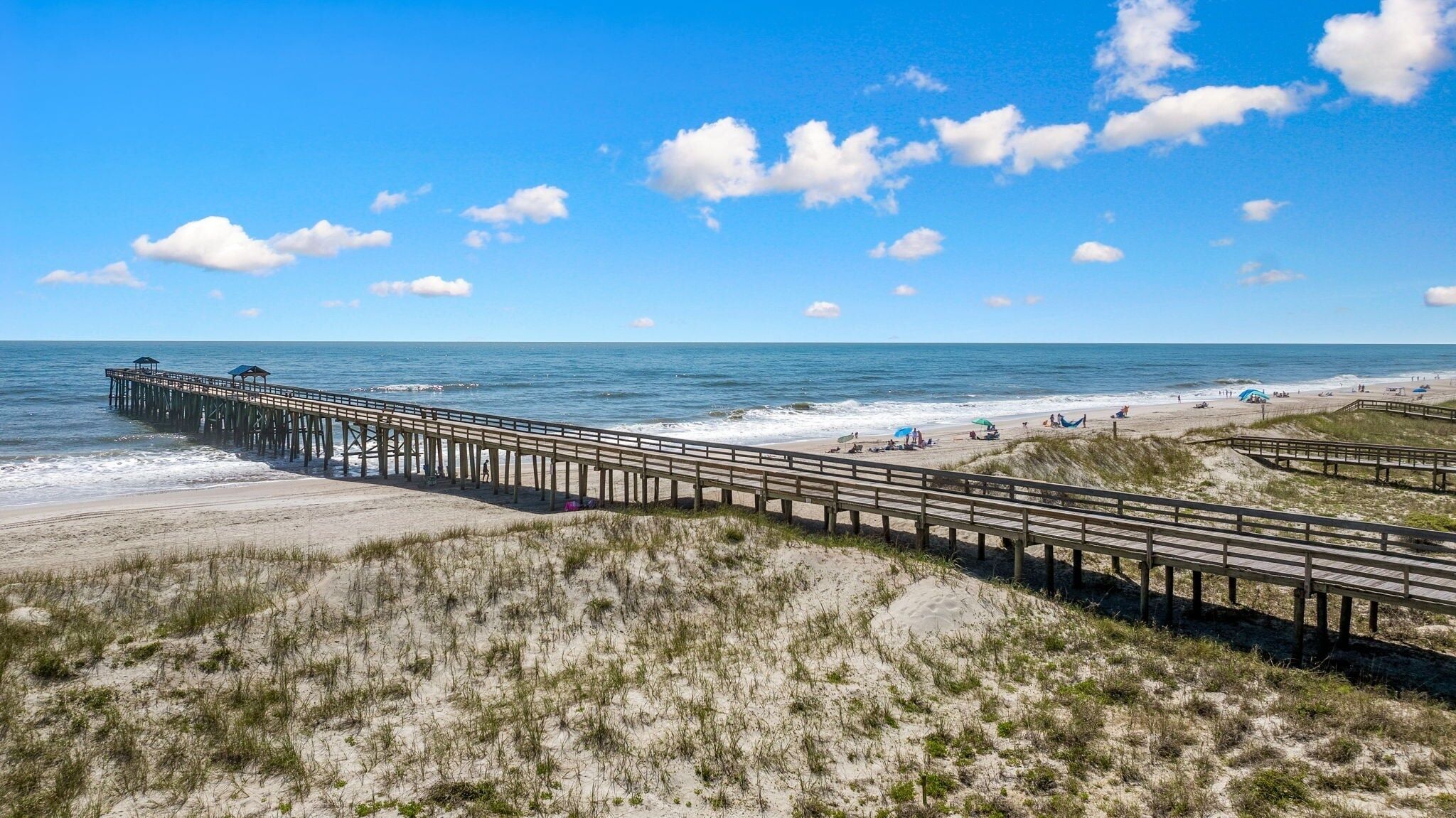 A beach with a wooden pier extending into the ocean, sandy dunes, and clear skies.