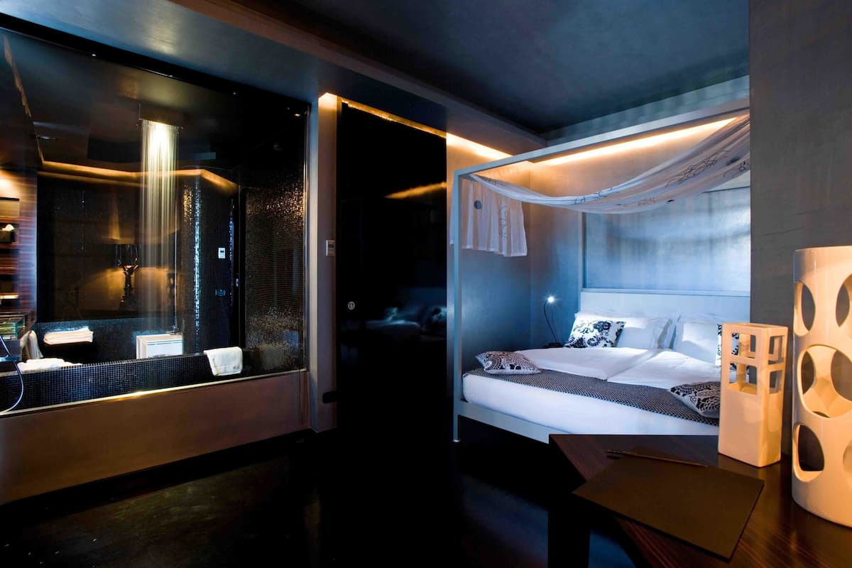 A modern hotel room with a large mirror, a bed with white linens, a bedside table, and a unique white decorative piece.