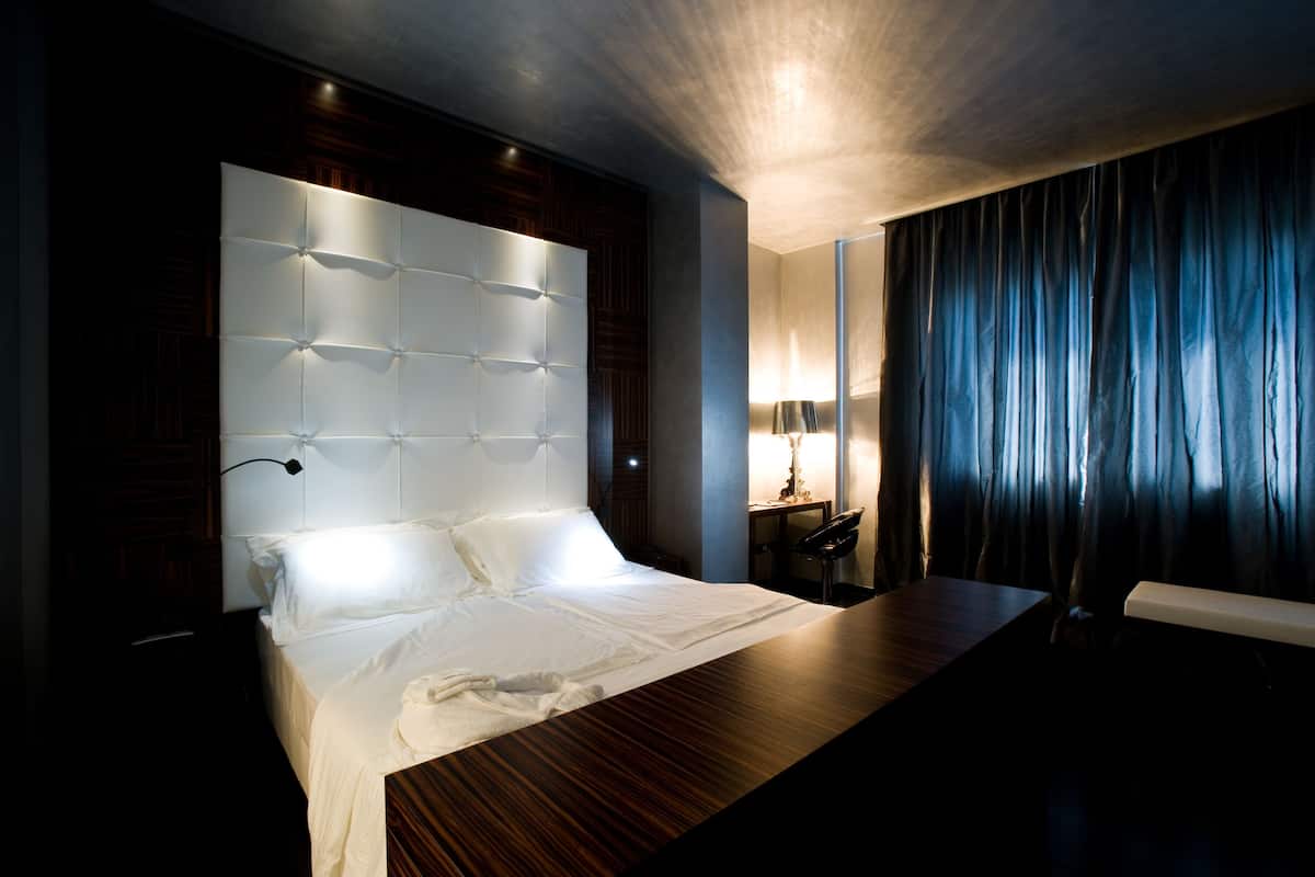 A modern hotel room with a large bed, a wooden desk, and a dark curtain backdrop.