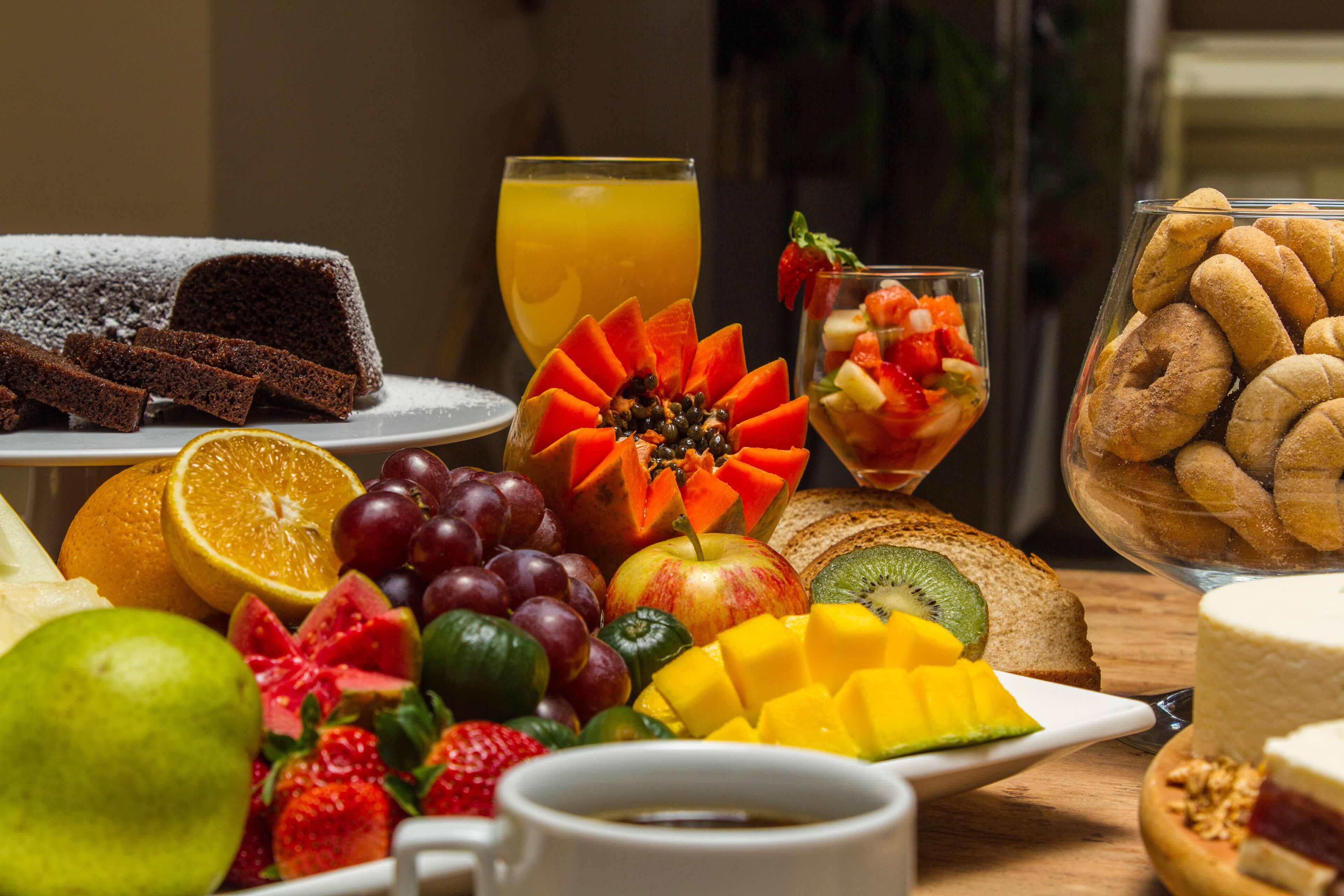 A breakfast table with various fruits, a glass of orange juice, a slice of cake, and a bowl of pastries.