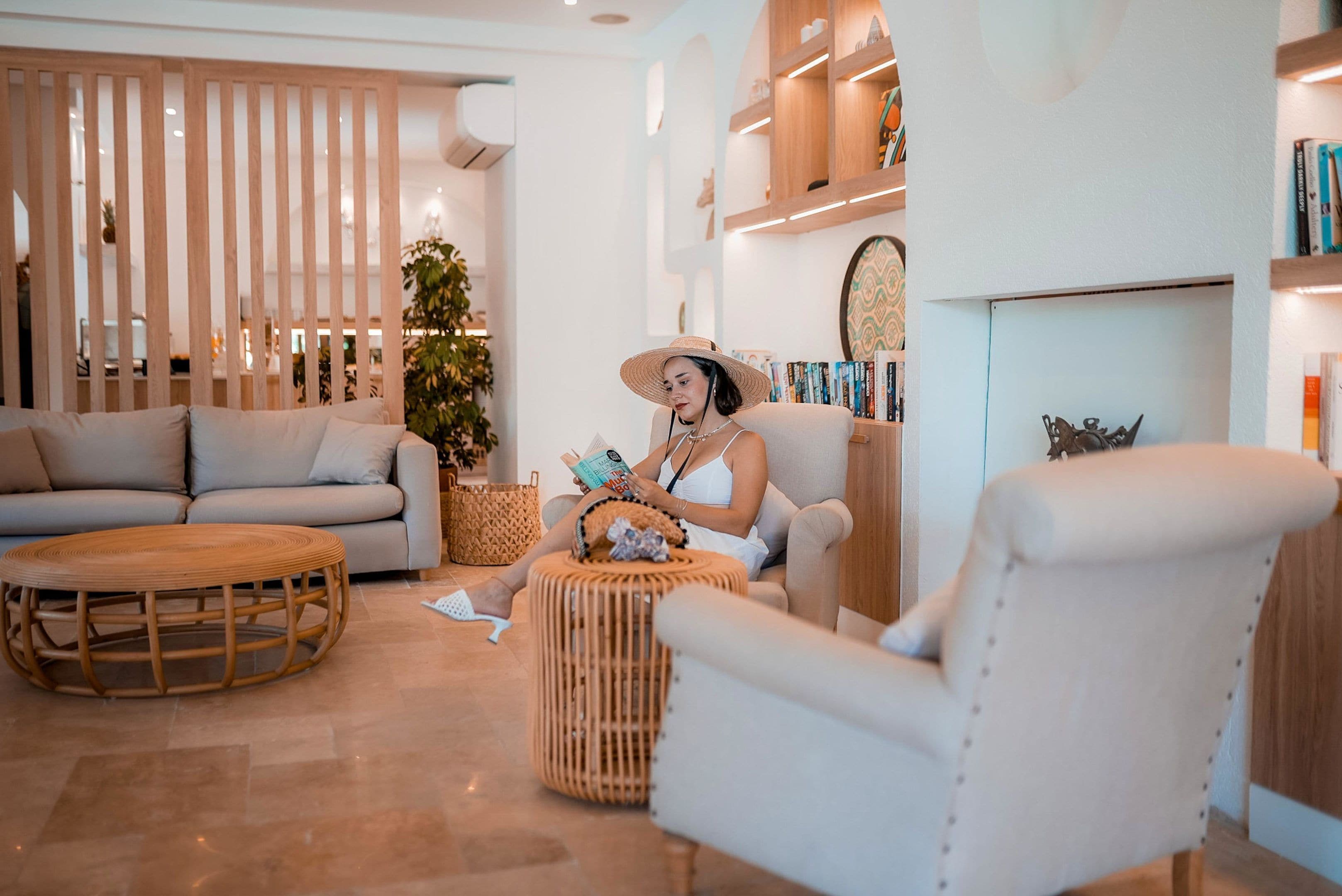Uma sala de estar moderna com sofá, poltrona e mesa de centro redonda de madeira. Há uma estante com livros e objetos decorativos.