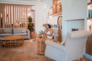Un salón moderno con un sofá, un sillón y una mesa de centro redonda de madera. Se observa una estantería con libros y objetos decorativos.