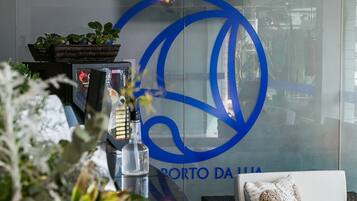 Un espace d'accueil avec un canapé en osier, un mur en verre orné d'un logo et des plantes en pot.
