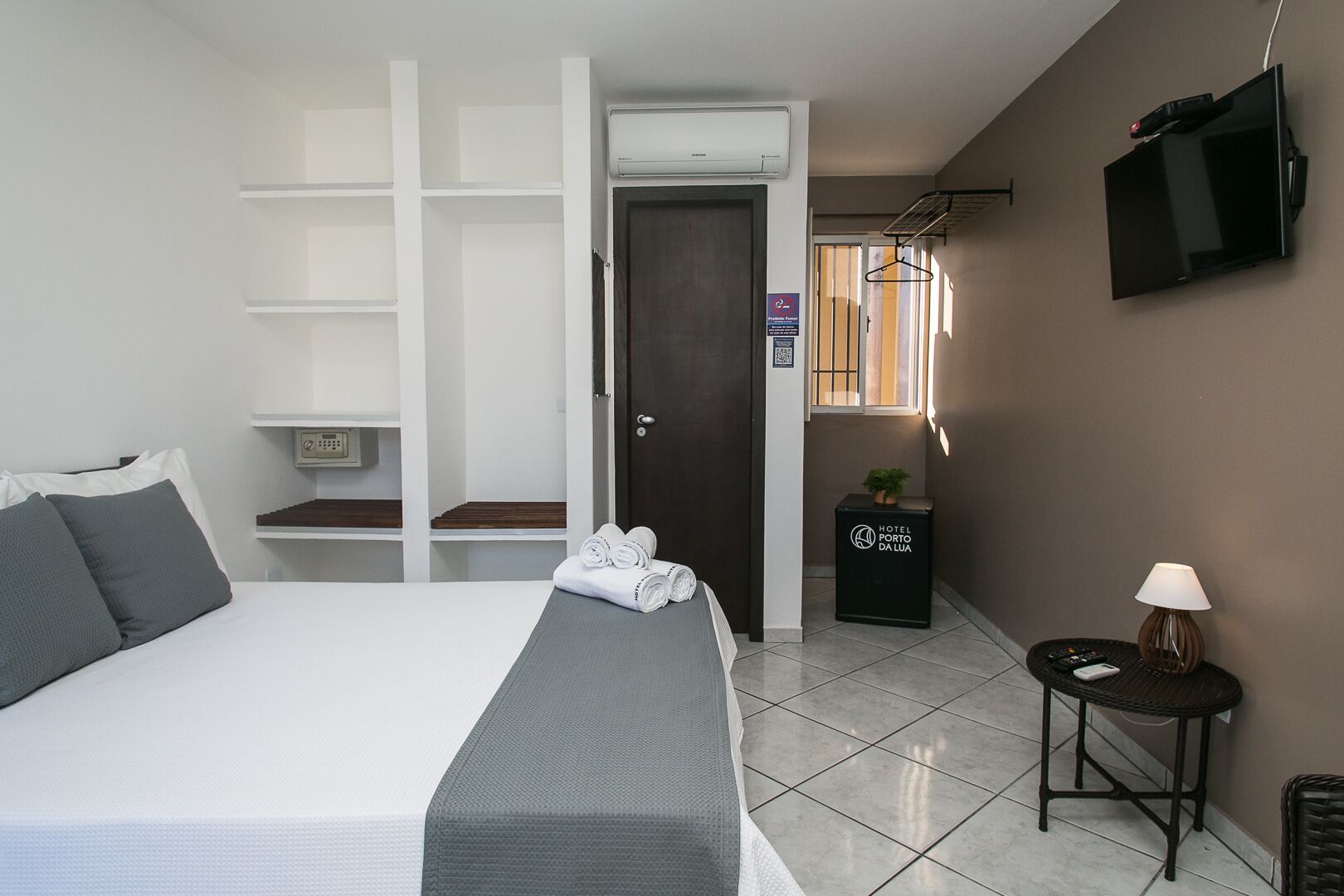 Una habitación de hotel con una cama, televisión, una mesita con lámpara, una planta y un cartel con el nombre del hotel.