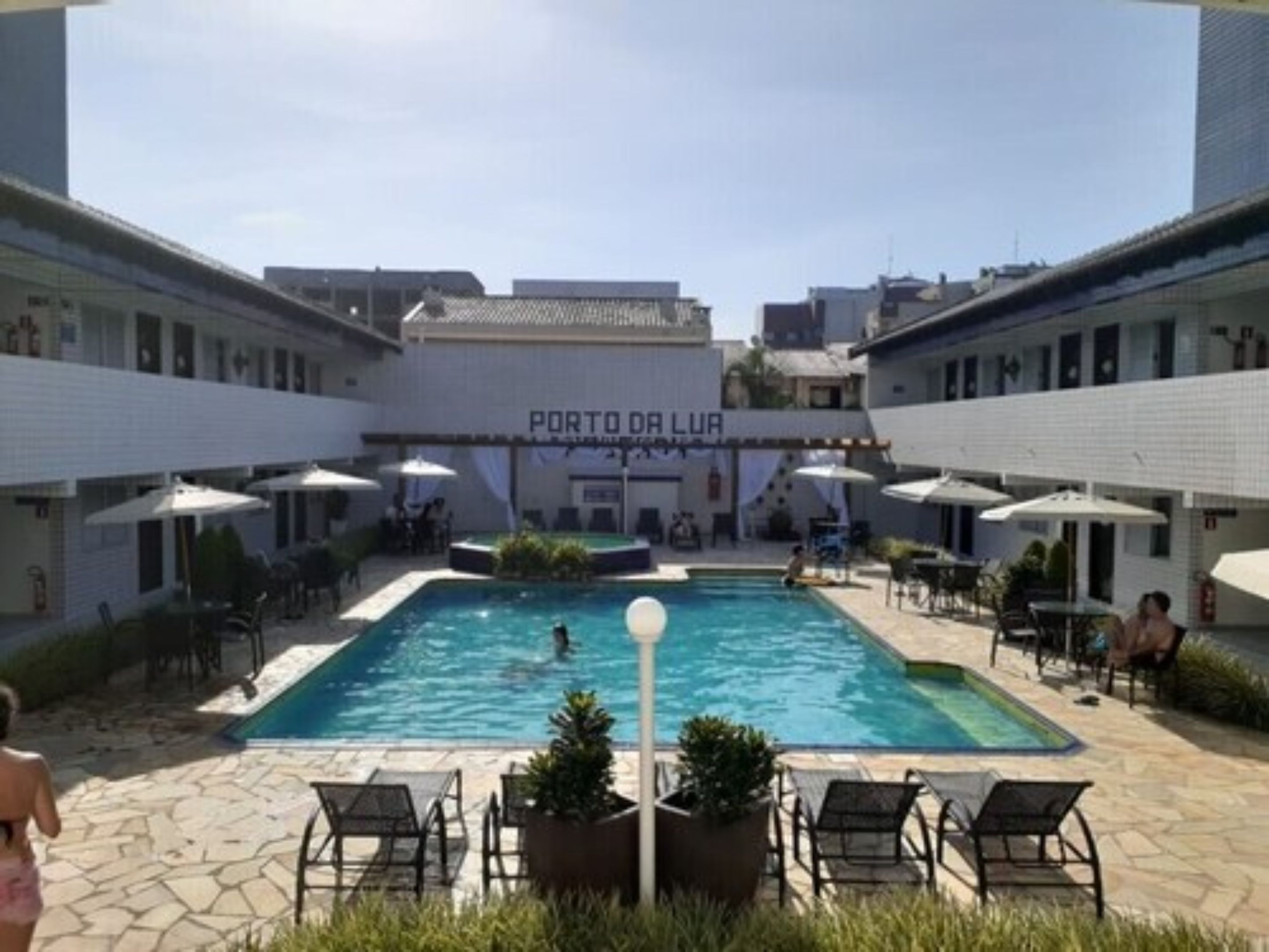 Zona de piscina con sillas de descanso y sombrillas, rodeada de edificios con un cartel que dice 'PORTO DA LUR'.