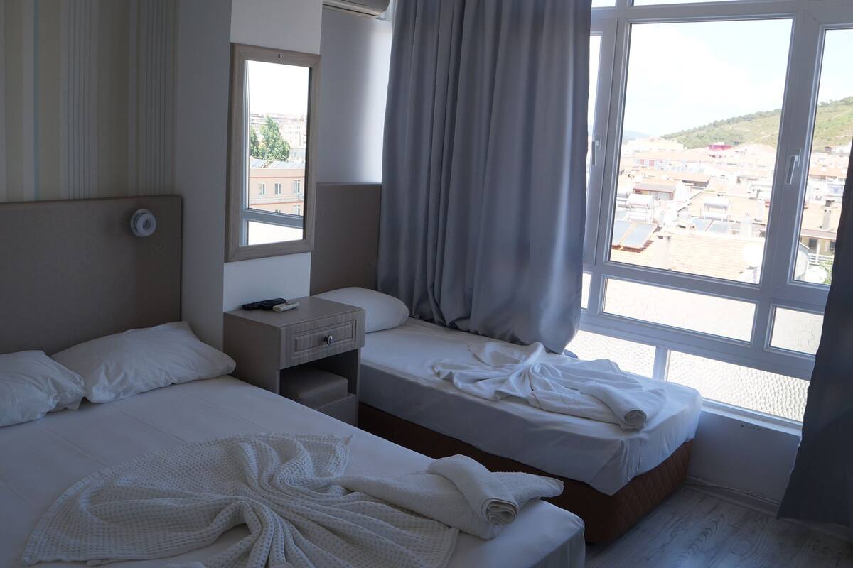 A hotel room with two beds, a nightstand, a window with curtains, and a view of buildings outside.