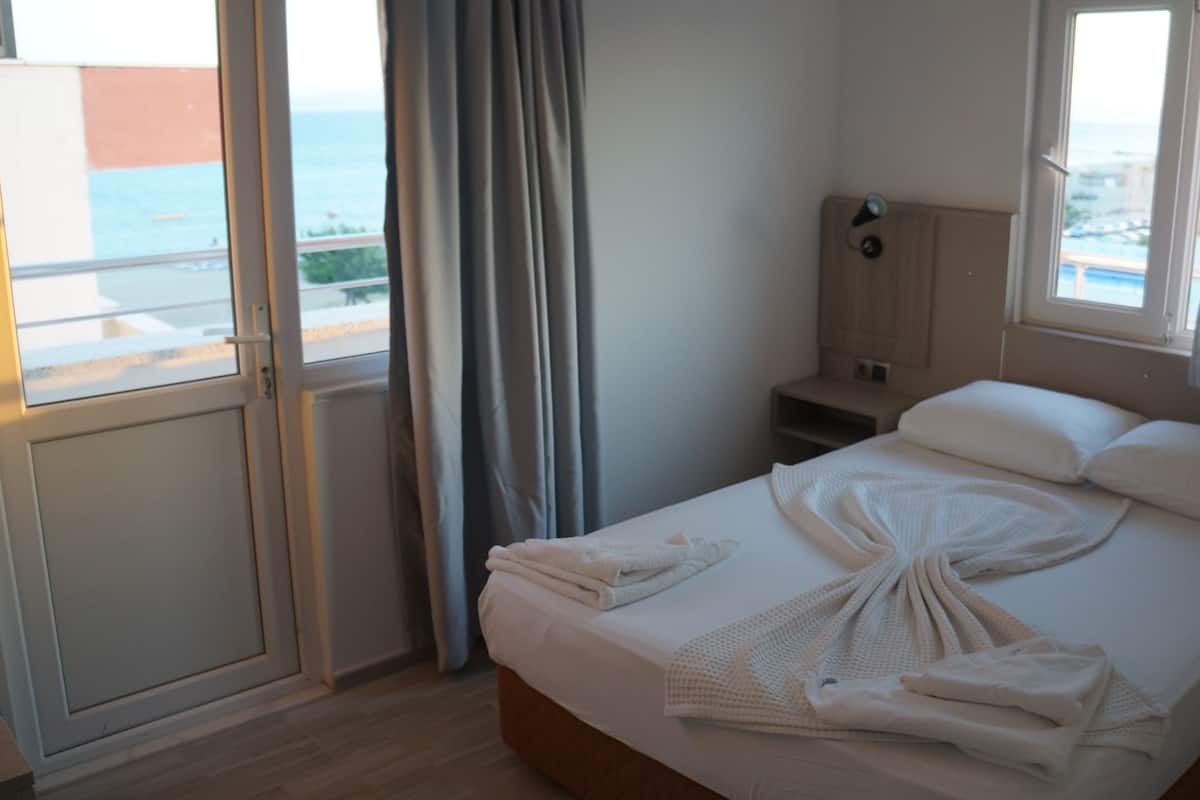 A hotel room with a bed, a nightstand, a lamp, and a view of the sea through the window.