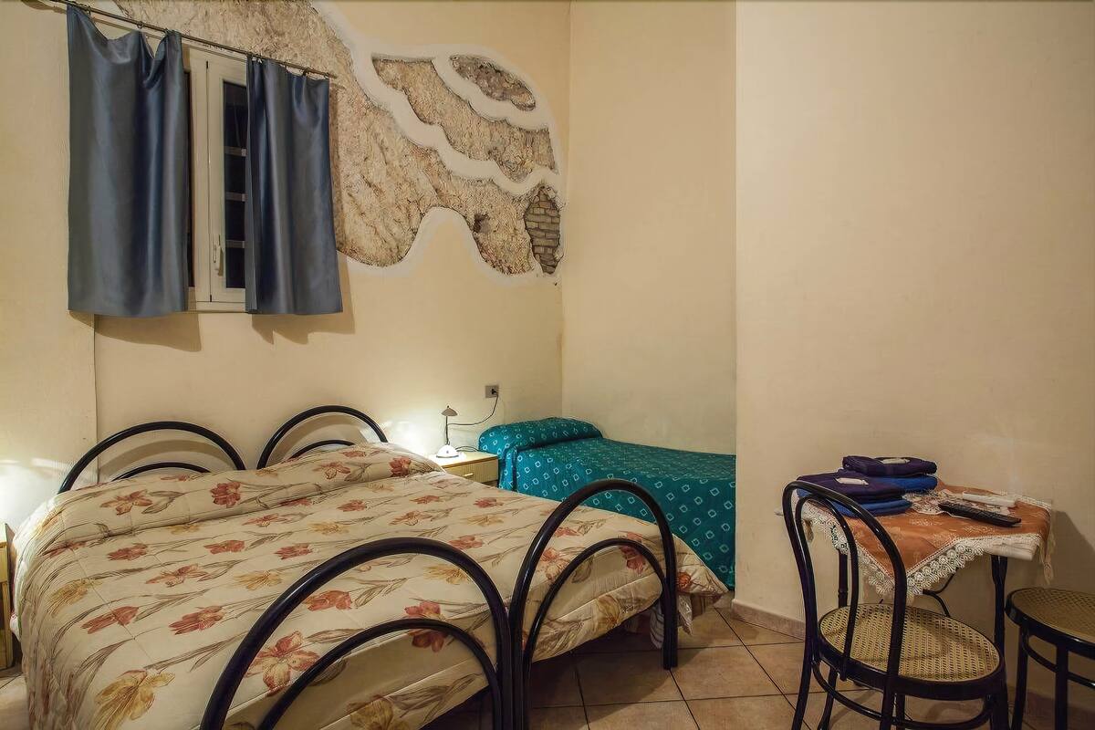 Un dormitorio con cama, una mesita, una silla y una ventana con cortinas.