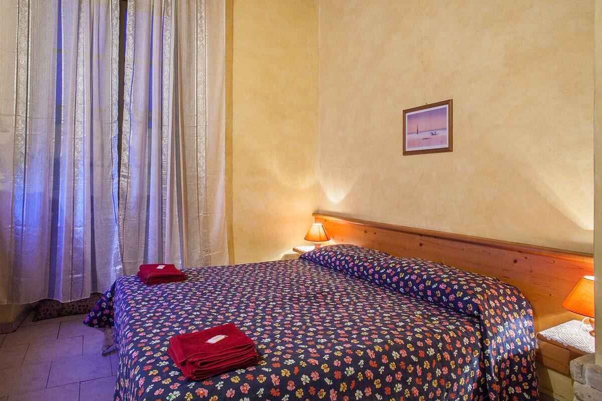 Habitación de hotel con cama, mesita de noche con lámpara, televisor sobre una mesa de madera y ventana con cortinas.