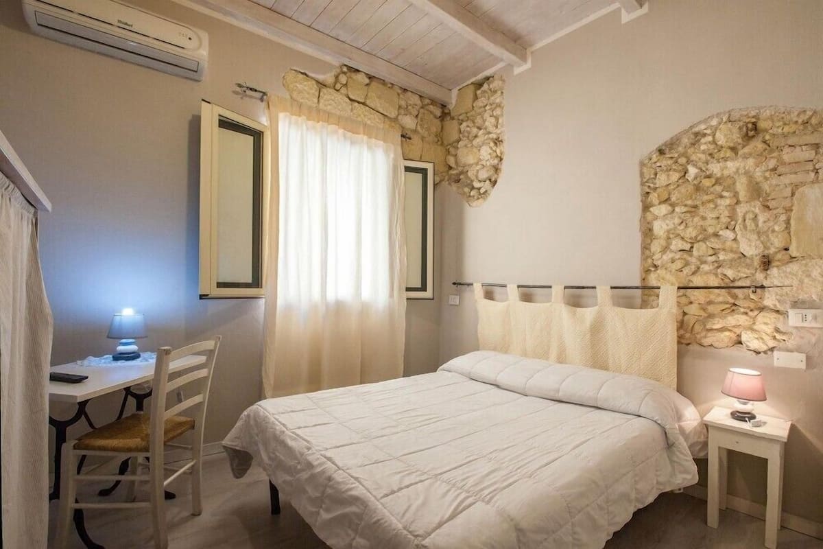 Un dormitorio con cama, un escritorio con lámpara, una silla, pared de piedra y una ventana con cortinas.