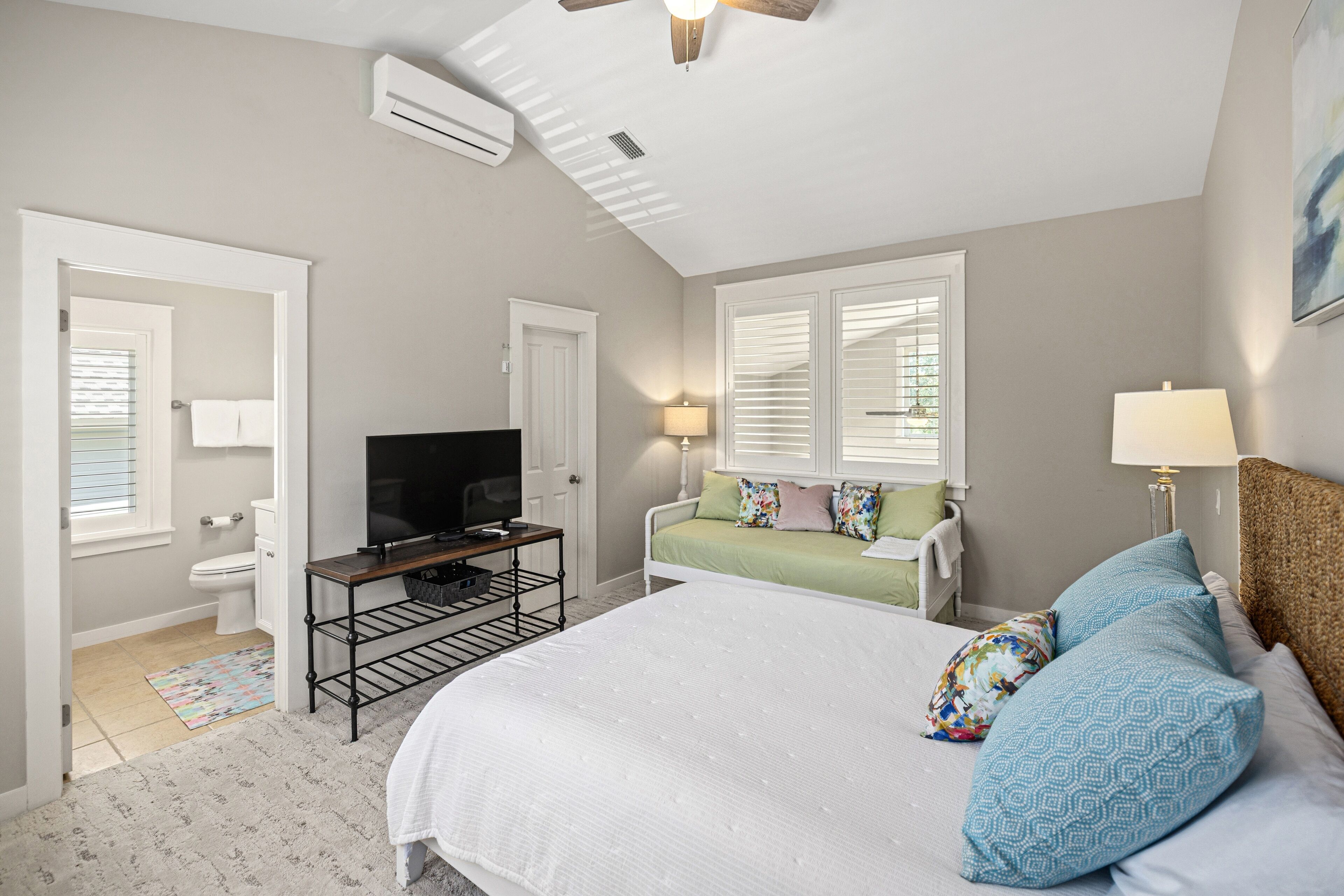 A bedroom with a bed, a television on a stand, a window with blinds, and a ceiling fan.