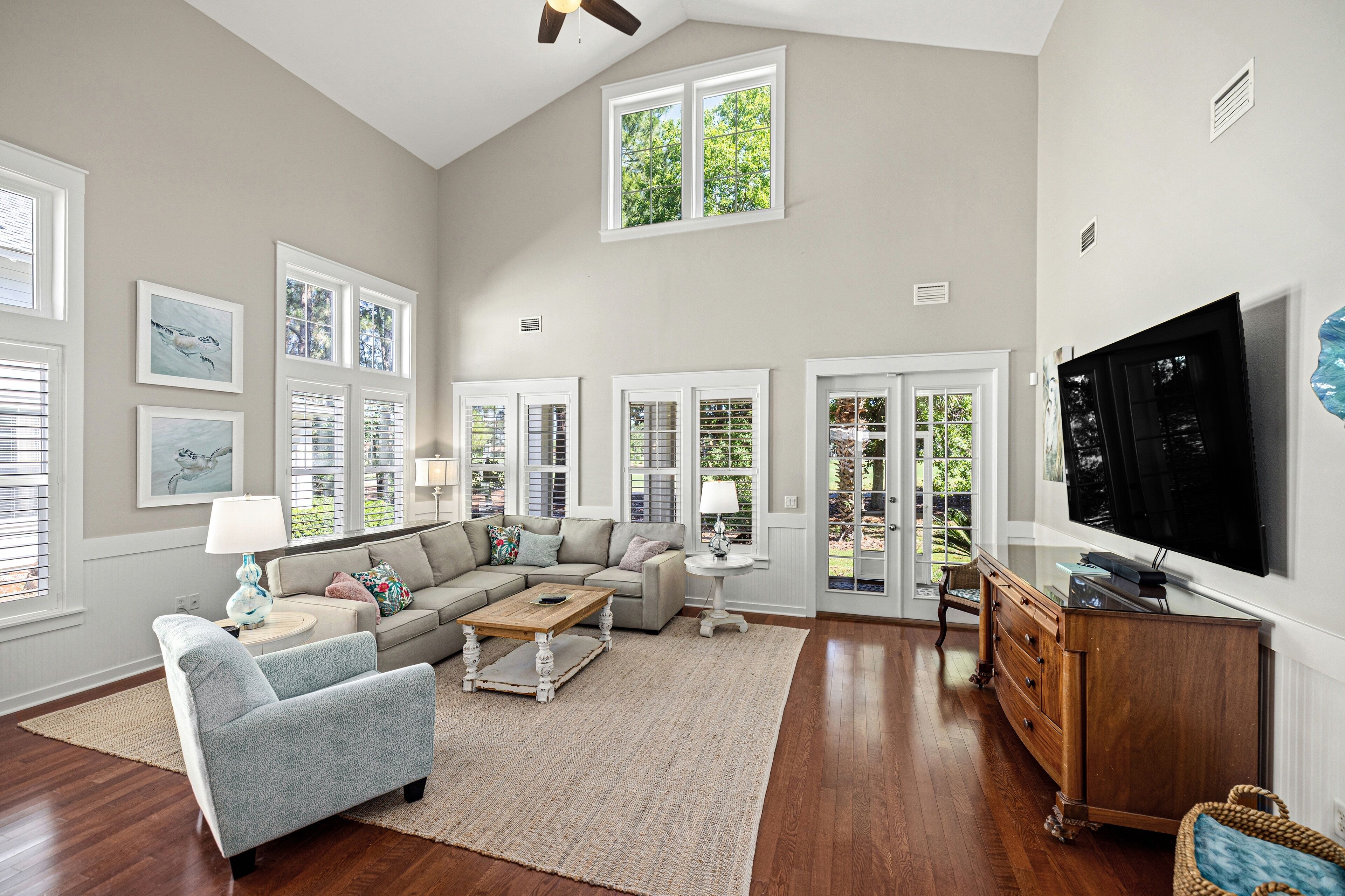 A spacious living room with a large sofa, a coffee table, and a television. There are multiple windows and a ceiling fan.