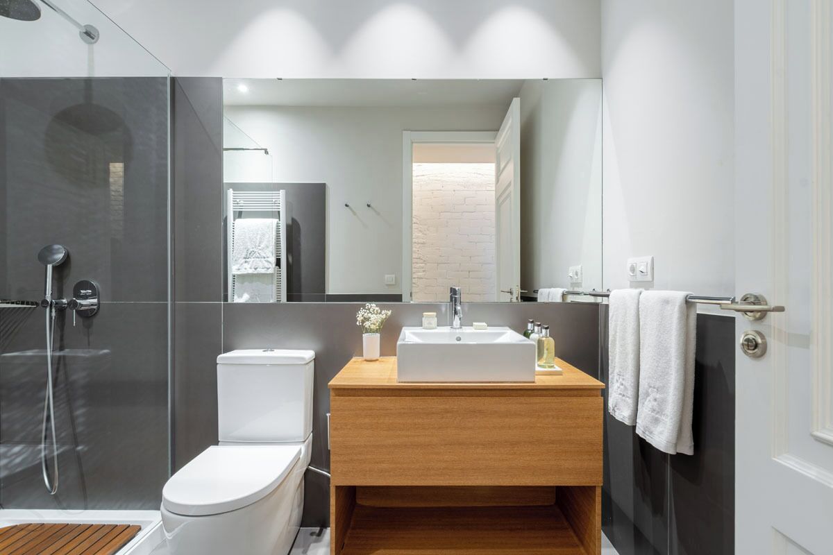 A modern bathroom with a glass shower enclosure, a white toilet, a wooden vanity with a sink, and a large mirror.